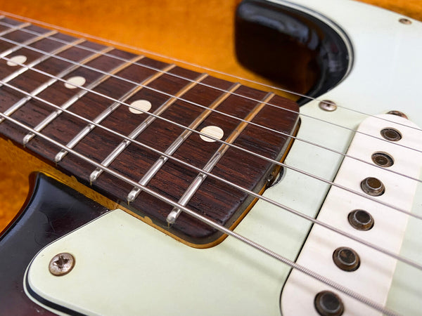 Close-up of electric guitar fretboard with metal strings, white pickguard, and vintage pickups on a sunburst body.