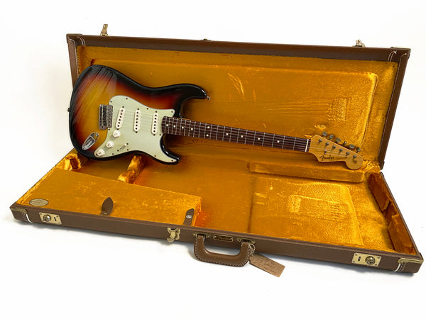 Vintage Fender Stratocaster electric guitar with sunburst finish resting in open brown hard case with plush orange interior lining