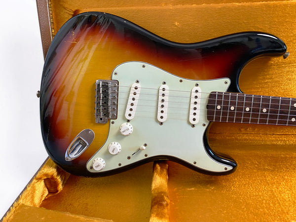 Close-up of vintage sunburst electric guitar with white pickguard, three single-coil pickups, volume and tone knobs, resting in yellow plush guitar case
