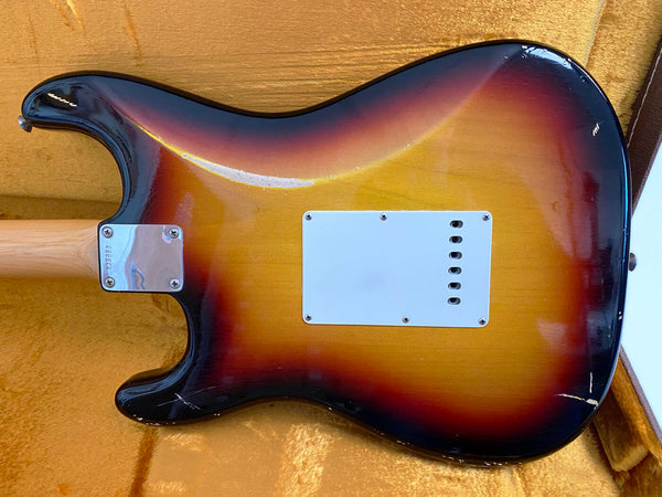 Back of sunburst electric guitar with white rectangular control plate and neck plate, showing worn finish and scratches, placed on yellow guitar case interior.