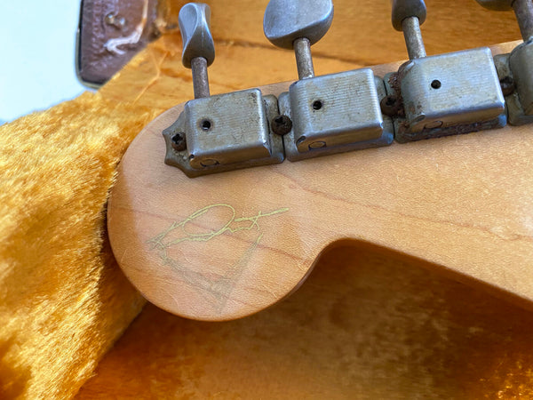 Close-up of vintage guitar headstock with metal tuning pegs and faded signature on maple wood, inside plush orange guitar case lining