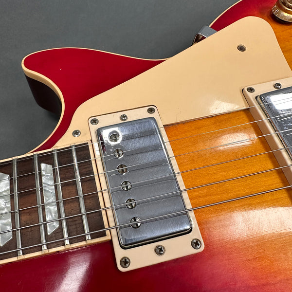 Close-up image of electric guitar body showing humbucker pickup, cream pickguard, rosewood fretboard with mother-of-pearl inlays, and sunburst finish on red and orange guitar top.