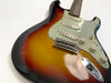 Close-up of vintage sunburst electric guitar body showing three single-coil pickups, volume and tone control knobs, and chrome bridge on white background.