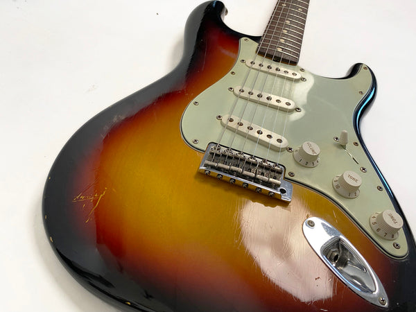 Close-up of vintage sunburst electric guitar body showing three single-coil pickups, volume and tone control knobs, and chrome bridge on white background.
