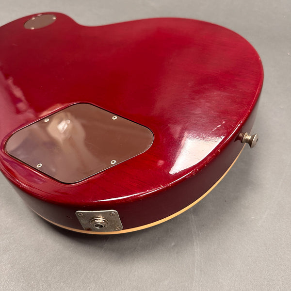 Close-up of the back of a red electric guitar body showing rear control cavity cover and jack plate with strap button on gray surface