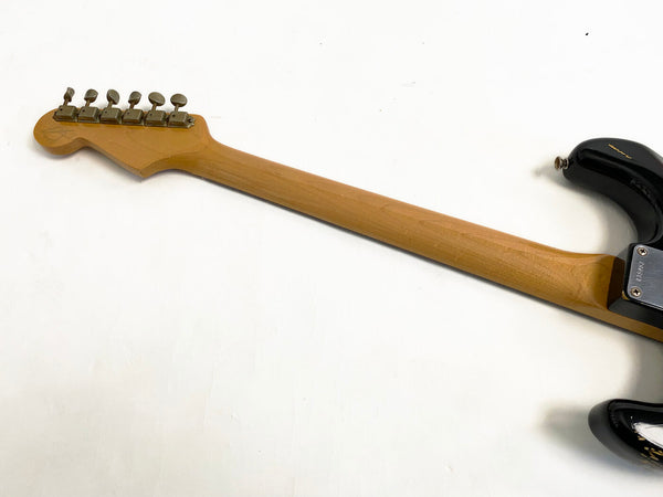 Back view of electric guitar neck with six tuning pegs, natural wood finish on the neck, and black body partially visible against white background