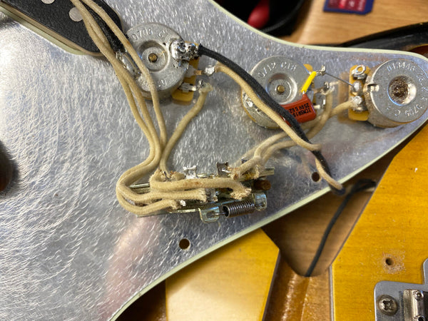 Close-up of electric guitar internal wiring and potentiometers on a silver pickguard with vintage cloth-covered wires and yellow capacitor, showing the tone control circuit details.
