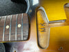 Close-up of guitar neck and fretboard with dot inlays next to the guitar body cavity with initials "MK" carved inside, showing sunburst finish and wood grain details.