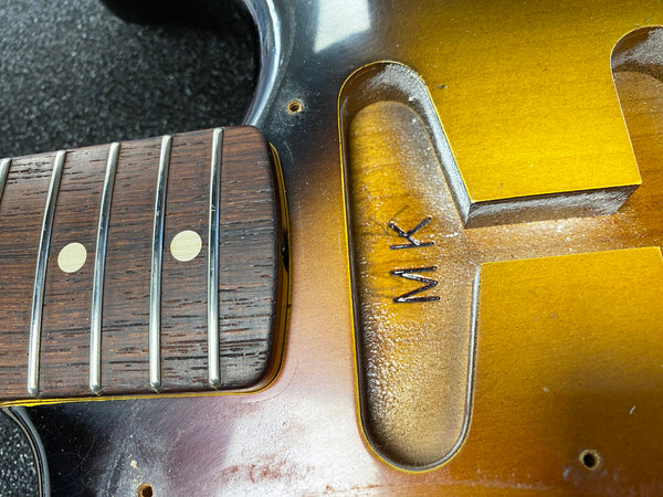 Close-up of guitar neck and fretboard with dot inlays next to the guitar body cavity with initials "MK" carved inside, showing sunburst finish and wood grain details.