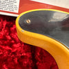 Close-up of vintage guitar body with yellow finish and black pickguard showing wear and checking, resting on red plush fabric background