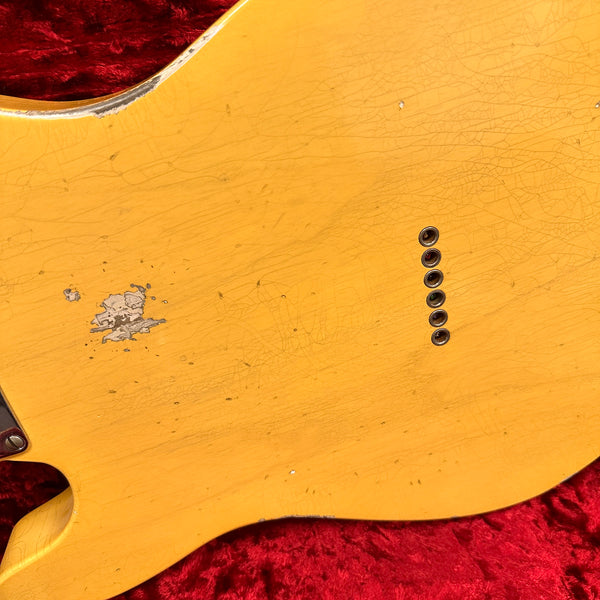 Close-up of vintage yellow electric guitar body with worn and chipped finish, showing string ferrules and fine checking, on red plush background