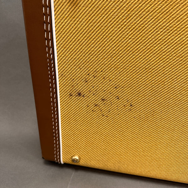 Close-up view of tweed guitar amplifier corner with brown leather trim and white piping, showing stitching details and a metal screw on gray background