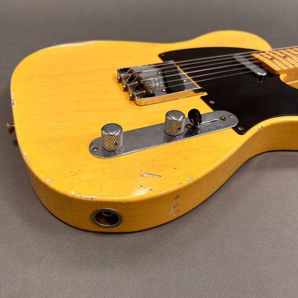 Close-up of vintage yellow electric guitar body with metal control knobs, black pickguard, worn finish, and chrome bridge on gray background