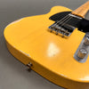 Close-up of vintage yellow electric guitar body showing wear and aged finish, black pickguard, chrome bridge, and control knobs on gray background.