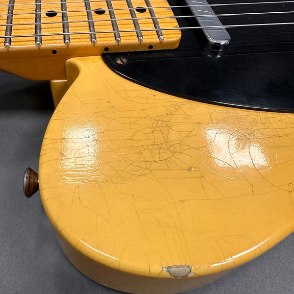 Close-up of vintage yellow electric guitar body with visible wear, cracked finish, and black pickguard next to fretboard with metal strings and inlay dots.