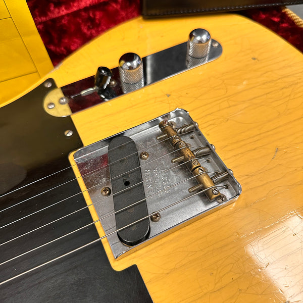Close-up of vintage Fender electric guitar bridge, pickup, and control knobs on worn yellow body with black pickguard and visible string tension adjustments