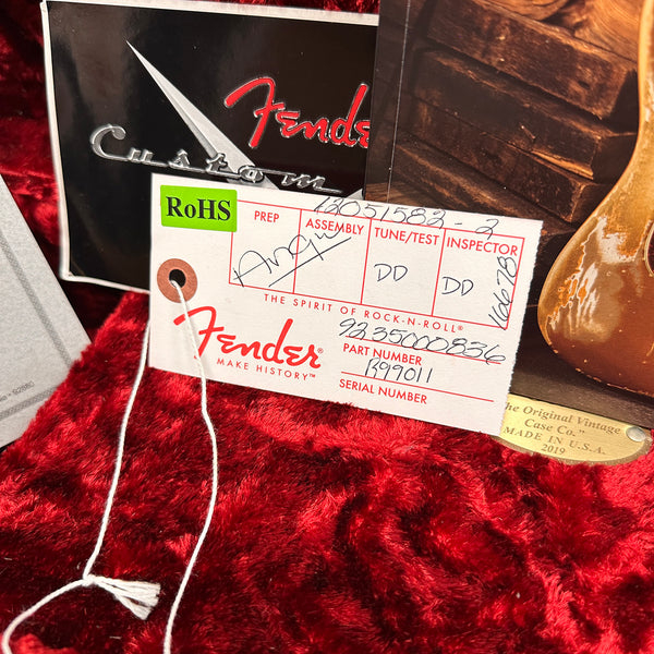 Close-up of Fender Custom Shop guitar tag with part number B99011 and serial number, placed on red plush guitar case interior with a partial view of the guitar body and vintage case label.