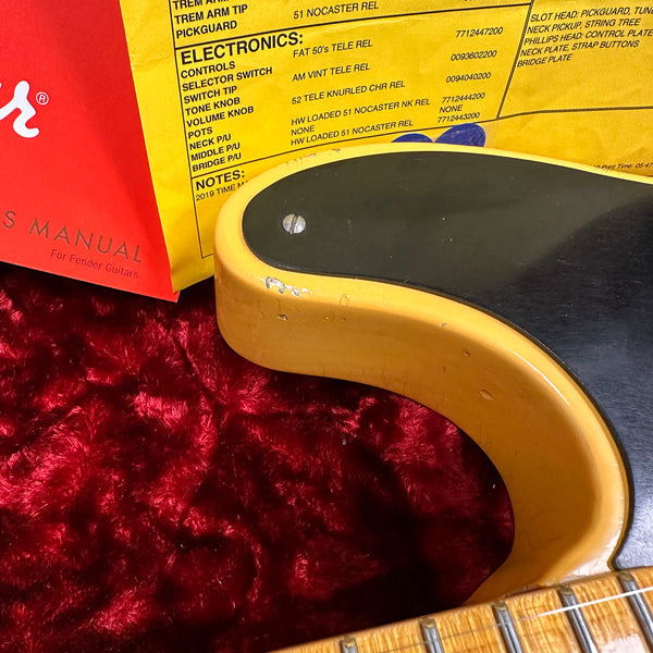 Close-up of a vintage-style Fender guitar body corner with black pickguard, yellow finish, visible wear, set against red plush fabric background, accompanied by Fender electronics specification sheet and manual for Fender guitars.