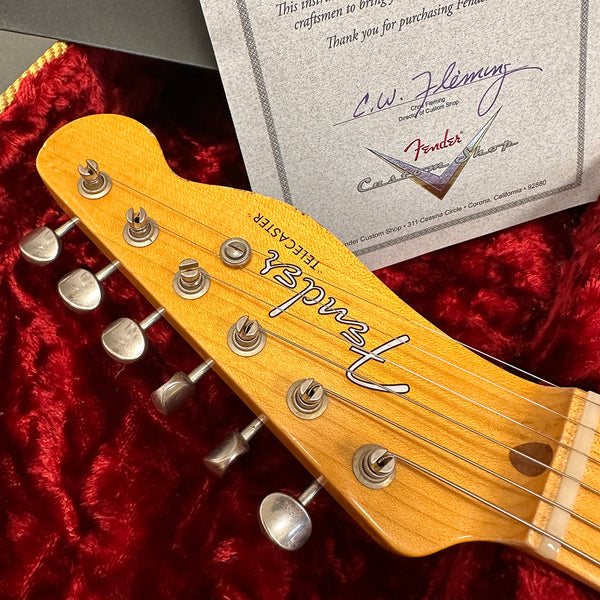Close-up of Fender Telecaster guitar headstock with tuning pegs, wood grain visible, resting on red velvet fabric with Fender Custom Shop certificate in the background