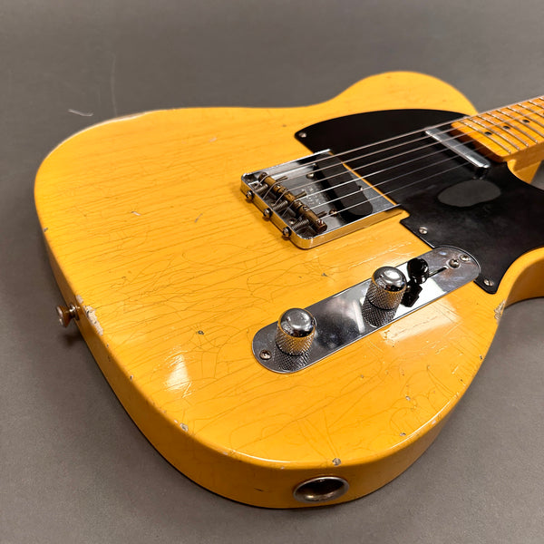 Close-up of vintage yellow electric guitar body with black pickguard, chrome bridge, and control knobs on gray background.