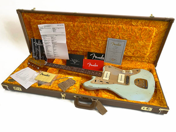 Fender vintage-style electric guitar in light blue finish with visible wear and relic relicing, shown inside a brown hard case lined with bright orange plush interior, accompanied by Fender certificates, manuals, cleaning cloth, and dealer spec sheet