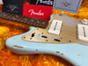 Close-up of vintage Fender electric guitar body and neck with worn light blue finish, white pickups, gold pickguard, and Fender manuals displayed on orange plush guitar case lining.
