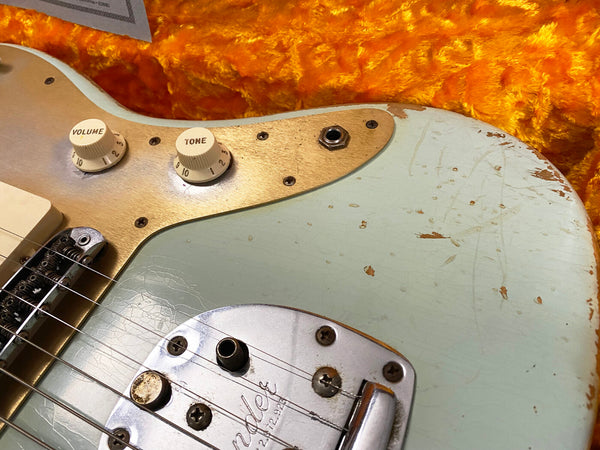 Close-up of vintage Fender electric guitar body with volume and tone knobs, showing wear and relic finish on light blue paint and gold pickguard against orange plush case lining