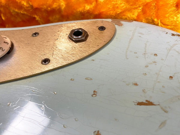 Close-up of worn vintage electric guitar body with aged cream pickguard, rusted input jack, and faded white finish showing paint chips and cracks against orange fabric background.