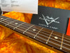 Close-up of Fender Custom Shop guitar fretboard with detailed metal frets and string winding, resting on orange plush lining with Fender Custom Care documentation in background.