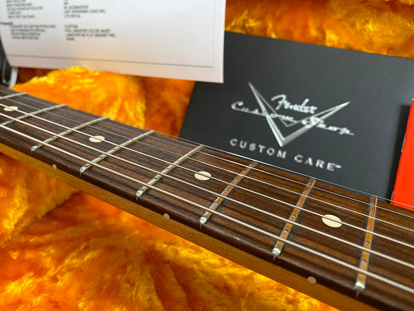 Close-up of Fender Custom Shop guitar fretboard with detailed metal frets and string winding, resting on orange plush lining with Fender Custom Care documentation in background.
