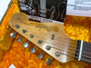 Close-up of Fender Jazzmaster guitar headstock with tuning pegs and strings, resting on orange plush case lining, with owner's manual in background.