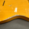 Close-up of yellow electric guitar body with visible paint cracks and worn area revealing wood underneath near the edge, showing vintage relic finish and string ferrules.