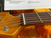 Close-up of guitar headstock and fretboard showing strings, nut, tuning peg, and part of a product specification sheet in the background with orange plush lining in guitar case.