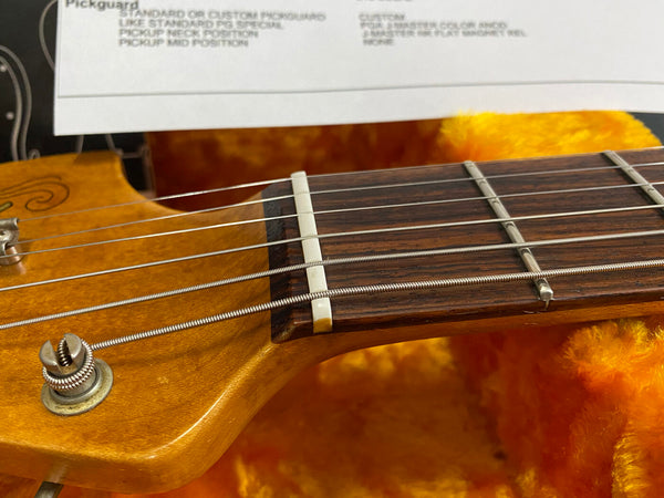 Close-up of guitar headstock and fretboard showing strings, nut, tuning peg, and part of a product specification sheet in the background with orange plush lining in guitar case.