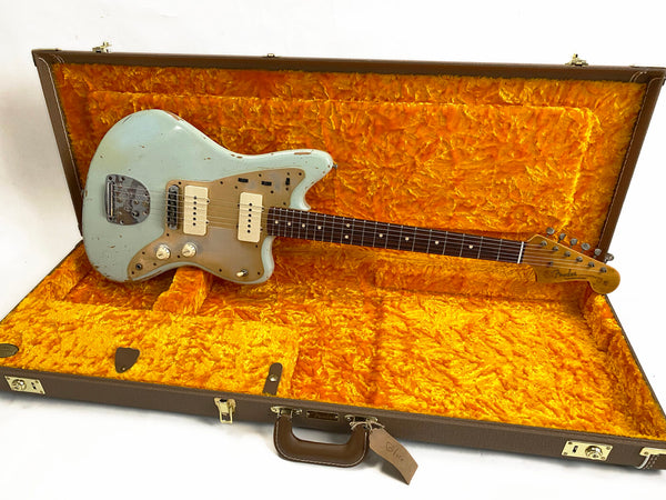 Vintage Fender Jazzmaster electric guitar in faded blue finish with rosewood fretboard, resting in open brown hard case with bright orange plush interior lining.