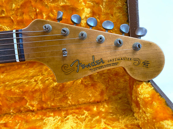 Close-up of Fender Jazzmaster guitar headstock with tuning pegs and strings, resting in a vintage plush orange guitar case