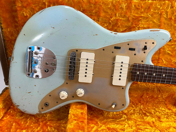 Vintage Fender Jazzmaster electric guitar in light blue finish with worn gold anodized aluminum pickguard, dual cream soapbar pickups, white control knobs, and rosewood fretboard on orange plush guitar case interior