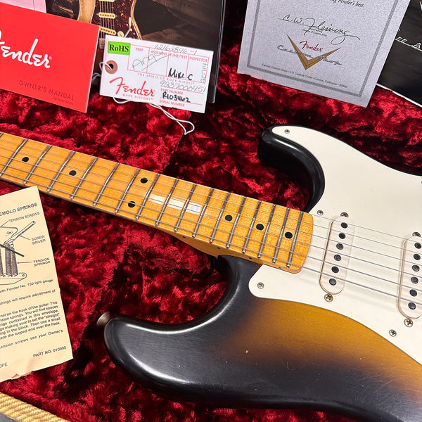Close-up of vintage Fender Stratocaster electric guitar neck and body with sunburst finish, surrounded by Fender owner's manual, tremolo springs instructions, and Fender Custom Shop certificates on red velvet guitar case lining.
