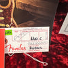 Close-up of a Fender guitar assembly tag with handwritten serial number R103462 and part number 9237000451 on a red textured fabric background.