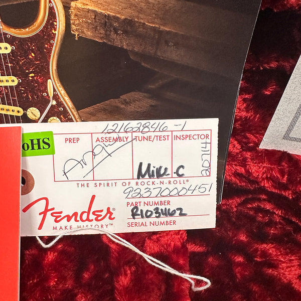 Close-up of a Fender guitar assembly tag with handwritten serial number R103462 and part number 9237000451 on a red textured fabric background.