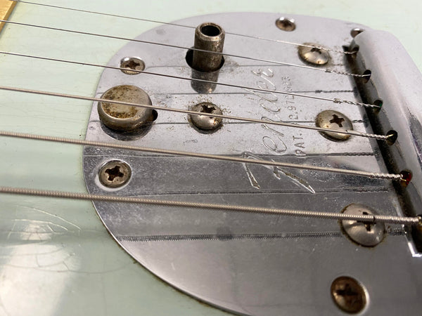 Close-up of vintage Fender guitar bridge and strings on light green guitar body showing engraved Fender logo and screws