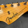 Close-up of Fender Stratocaster headstock with tuning pegs and strings, featuring natural wood finish and logo with synchronized tremolo text.