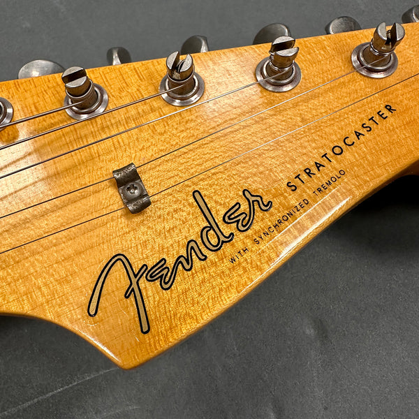 Close-up of Fender Stratocaster headstock with tuning pegs and strings, featuring natural wood finish and logo with synchronized tremolo text.