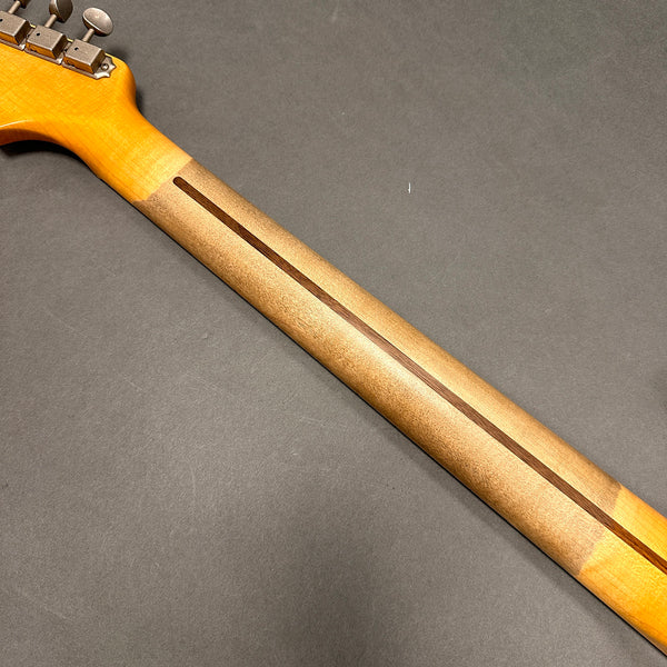 Close-up of a guitar neck showing the hardwood construction with a dark wood reinforcement strip along the back and vintage-style tuning pegs against a gray background.