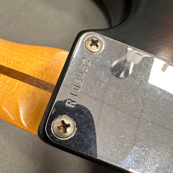 Close-up of guitar neck joint plate with serial number R103462 and two screws on a black electric guitar with a natural wood neck