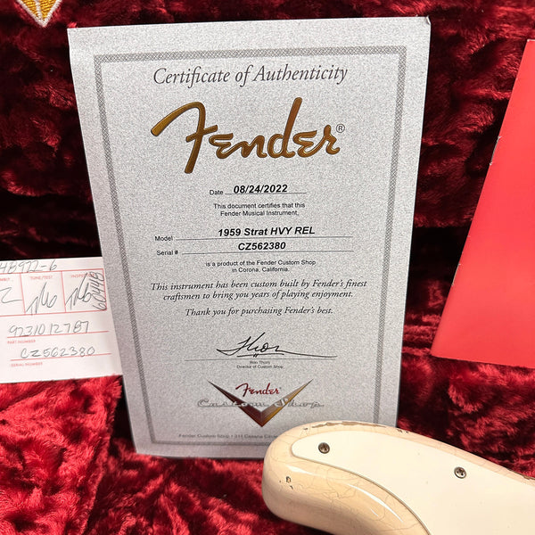 Fender Certificate of Authenticity for 1959 Strat HVY REL with serial number CZ562380, dated 08/24/2022, displayed on red plush fabric next to a guitar body and Fender Custom Shop paperwork.