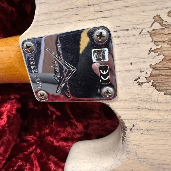 Close-up of Fender Custom Shop guitar neck plate with serial number and worn, cracked white finish on guitar body against red velvet background