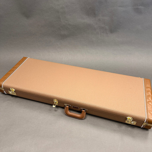 Closed tan hardshell guitar case with brown leather handle and gold latches on gray background