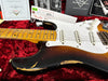 Close-up of a vintage Fender Stratocaster electric guitar with a worn sunburst finish, maple neck, and three single-coil pickups, resting on a red plush guitar case with Fender custom care documentation and inspection tags in the background.