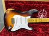 Vintage Fender Stratocaster electric guitar with sunburst finish and white pickguard, showing wear and aging, resting in red plush-lined hard case embroidered with "Stratocaster 60 Years 1954-2014" logo